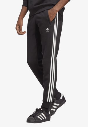Mand iført sorte Adidas træningsbukser med hvide striber og sorte Adidas sneakers med hvide striber, stående med den ene hånd i lommen.
