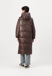 Abrigo acolchado marrón con capucha, con secciones acolchadas horizontales y cremalleras laterales, combinado con pantalones de mezclilla claros y zapatillas.