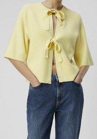 Cardigan giallo a coste con maniche corte, dotato di due fiocchi sul davanti, scollo aperto e design cropped abbinato a jeans di denim a vita alta.