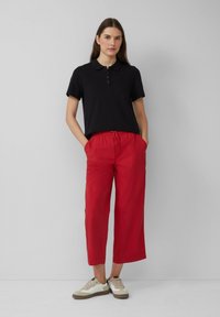 Polo noir à manches courtes avec une patte de boutonnage à trois boutons, associé à un pantalon rouge large et court, et des baskets beiges avec des accents noirs.