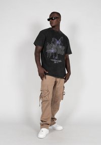 Sort grafisk T-shirt med sommerfugledesign, parret med beige cargo-bukser med sidelommer. Modellen har hvide sneakers og solbriller på.