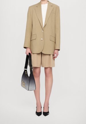 Beige blazer with two buttons and patch pockets, paired with beige shorts. Black handbag with a gradient effect and gold lettering. Black heels.