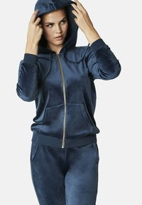 Haut de survêtement zippé en velours doux de couleur bleu marine, avec poches avant, poignets côtelés et capuche. Porté avec un pantalon en velours assorti.