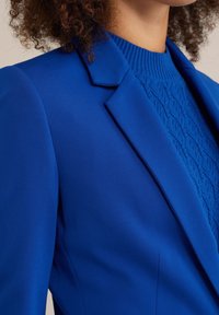 Blazer blu in tessuto liscio con rever a punta, abbinato a un maglione in maglia di cotone blu a trama. Vista ravvicinata che mette in evidenza i dettagli di colore e design.