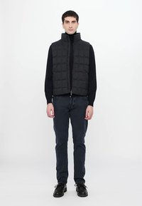 Black quilted vest with high neck, zip closure, and contrasting texture, worn over a black turtleneck and paired with dark jeans and shoes.