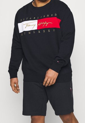 Homme portant un sweat-shirt noir Tommy Hilfiger avec un panneau rouge et blanc et des lettres dorées, assorti à un short noir avec un petit logo sur la jambe gauche.