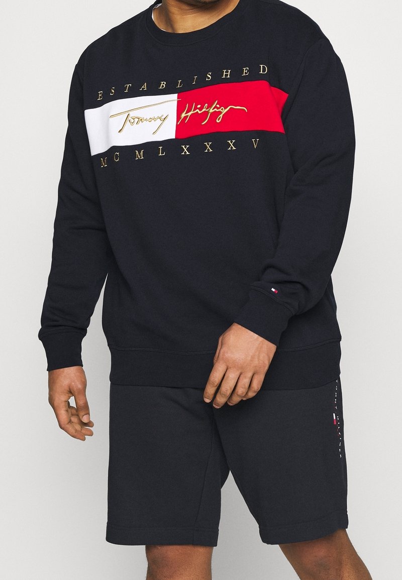 Homme portant un sweat-shirt noir Tommy Hilfiger avec un panneau rouge et blanc et des lettres dorées, assorti à un short noir avec un petit logo sur la jambe gauche.