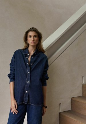 Jeune femme aux longs cheveux portant une chemise en denim bleu foncé et un pantalon, debout près d’un escalier en bois contre un mur beige.