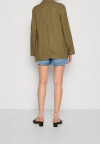 Olivgrön linnéblazer med avslappnad passform, långa ärmar och en ensam vent, i kombination med denimshorts och svarta klackmules.