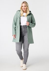 Lichtgroene waterdichte jas met een afneembare capuchon, zijzakken en een ritssluiting, gedragen over een wit shirt en grijze jeans.