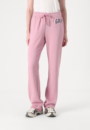 Personne portant un pantalon de jogging rose clair avec cordon de serrage à la taille et logo GAP sur la cuisse gauche, associé à des baskets blanches et noires.