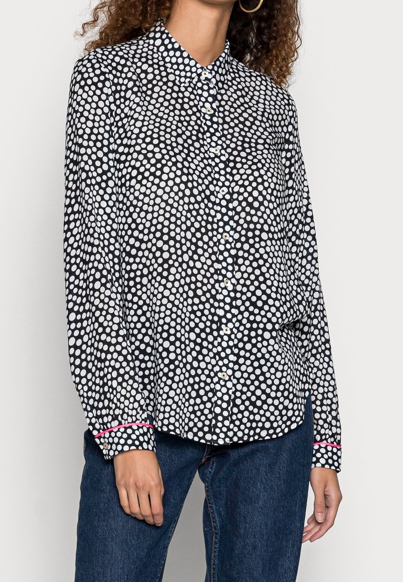 Femme portant une chemise à manches longues à pois noirs et blancs avec un bord rose aux poignets et un jean bleu, debout devant un fond blanc.