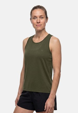 Femme à la peau claire avec les cheveux attachés, portant un haut sans manches vert olive et un short noir, debout devant un fond clair uni.