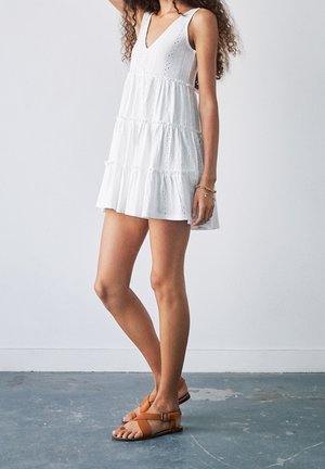 Robe blanche à étages avec un profond décolleté en V, détails en broderie anglaise et ourlet à volants ; portée avec des sandales brunes à lanières.