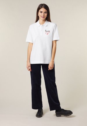 Camicetta polo bianca con un grafiche rossa "LOVE" e design di spilla da balia, abbinata a pantaloni a gamba larga in velluto a coste blu navy e scarpe nere.