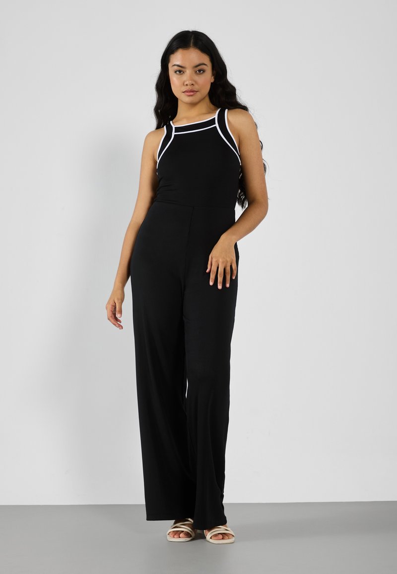 Combinaison noire avec un haut ajusté, bordures contrastées blanches sur les épaules et pantalons évasés. Texture lisse, design minimaliste.