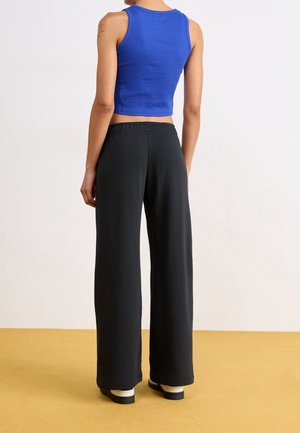 Person, der står iført en blå ærmeløs crop top og løse sorte bukser på en gul gulv med en simpel hvid baggrund.
