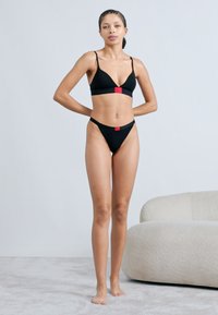 Black bikini set featuring a triangle bralette with thin straps and matching high-cut briefs. Red logo tag visible on the bralette.