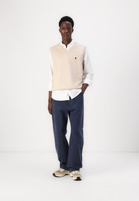 Μπεζ πλεκτή γιλέκα πάνω από ένα λευκό πουκάμισο με κουμπιά, σε συνδυασμό με χαλαρές σκούρες μπλε τζιν και ανοιχτόχρωμα αθλητικά παπούτσια. Απλός σχεδιασμός με μίνιμαλ λεπτομέρειες.