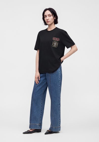 Γυναίκα όρθια φορώντας μαύρο γραφικό t-shirt, μπλε παντελόνι φαρδύ και μαύρα μυτερά μπαλαρίνια σε απλό φωτεινό φόντο.