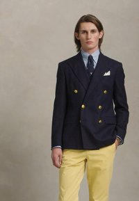 Tamno plavi dvoredni sako sa zlatnim dugmadi, prugasta plava i bijela košulja, žute hlače i bijeli rupčić.