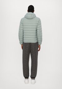 Lysegrøn pufferjakke med hætte, ribbede manchetter, kombineret med sorte mønstrede sweatpants og hvide sneakers, set bagfra.