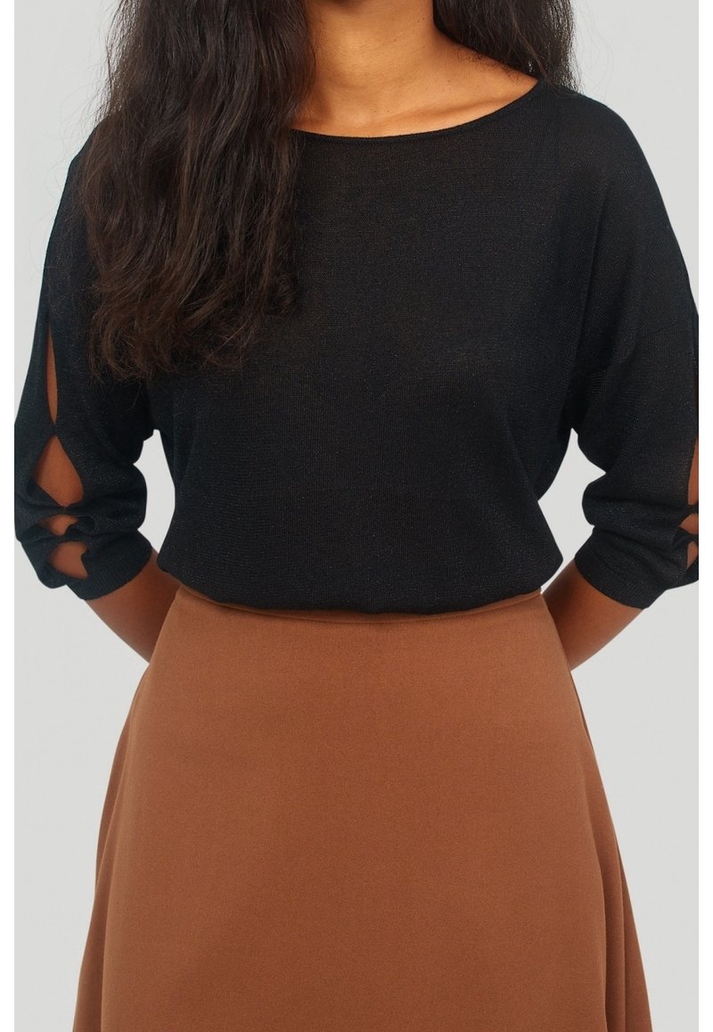 Femme portant un haut noir à manches longues avec des découpes sur les côtés et une jupe marron taille haute, debout devant un fond uni.