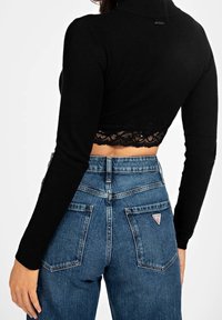 Černý, dlouhý crop top s krajkovým lemem ve spodní části, spárovaný s modrými džíny s vysokým pasem, které mají dvojité zadní kapsy a akcent s logem.