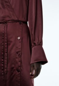 Bordeauxfarbiges Satin-Kleid mit Wickeldesign, langen Ärmeln, großen Glockenärmeln und runden Knöpfen entlang der Taille, die eine glatte Textur schaffen.