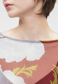 Διαφανής μπλούζα με λουλουδάτα σχέδια σε λευκό, χρυσό και ανοιχτό ροζ. Φαρδύ λαιμόκοψη με αντίθετη κίτρινη υφή και λεπτό δίχτυ.