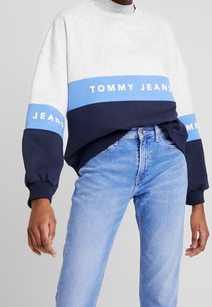 Persona con sudadera a bloques de color blanco, azul y azul marino con texto "Tommy Jeans", combinada con vaqueros azul claro.