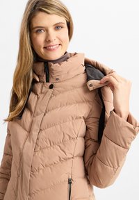 Hellbrauner Puffer-Jacke mit Chevron-Stich, hohem Kragen, zwei seitlichen Reißverschluss-Taschen und schwarzen Akzenten am inneren Kragen und den Schultergurten.