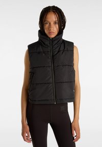 Svart pufferväst med hög krage, framdragkedja och kviltad design. Bärs över svarta åtsittande leggings med framträdande sömnadsdetaljer.