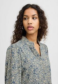 Blusa blu con un motivo in pizzo floreale beige, abbottonatura frontale e spalle plissettate. Il tessuto sembra leggero e testurizzato.