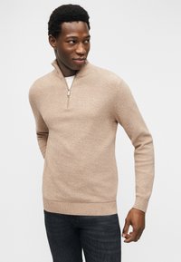 Άνδρας με μπεζ πουλόβερ με μισή φερμουάρ και σκούρο τζιν, κοιτώντας προς τα αριστερά του, με απλό λευκό φόντο.