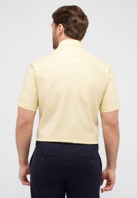 Camicia a maniche corte con colletto in tessuto giallo chiaro con un motivo testurizzato, abbinata a pantaloni scuri navy con tasche posteriori e dettagli con bottoni.