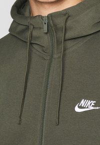 Nike zip-up hoodie i mörk olivgrön, med en mjuk textur, två dragkedjor och en vit logotypaccent på bröstet.