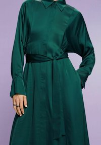 Robe en satin vert émeraude avec col, devant boutonné, taille ceinturée et manches longues avec détails aux poignets. Texture lisse et fluide.