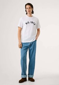 Hvit t-skjorte med korte ermer og svart "PJL 1973"-tekst, kombinert med blå jeans i denim og brune ankelstøvler. Enkel, avslappet design.