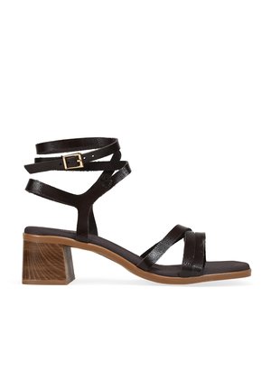 Sandalia negra con tiras y hebilla dorada, correa envolvente en el tobillo, tacón bloque de madera y suela marrón, vista de lado sobre fondo blanco.