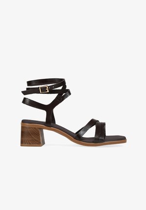 Sandalia negra con tiras y hebilla dorada, correa envolvente en el tobillo, tacón bloque de madera y suela marrón, vista de lado sobre fondo blanco.