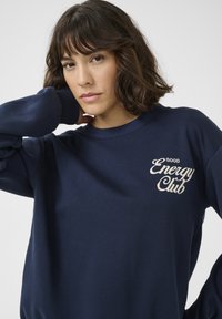 Tamnoplava dukserica napravljena od mekanog materijala. Ima okrugli izrez i rebraste manžete. "Good Energy Club" izvezena je svijetlom krem bojom na prsima.