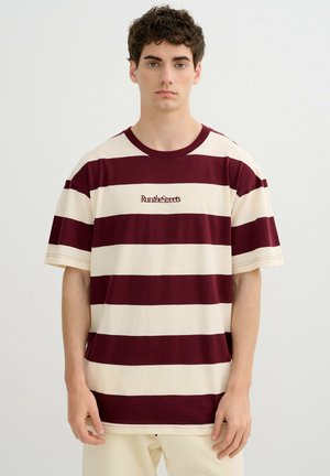 Hombre joven que lleva una camiseta de rayas horizontales en granate y crema con el texto "Run the Streets" en la parte delantera, de pie frente a un fondo liso.