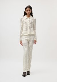 Háčkovaný bílý cardigan s límečkovým výstřihem a zapínáním na knoflíky, doplněný bílými ribbed kalhotami se vlnkovými vzory. Hnědé boty.