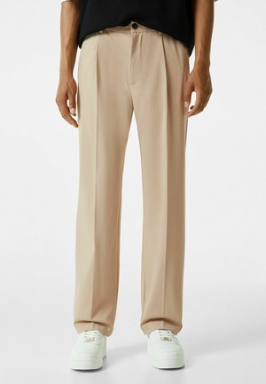 Man die beige rechte broek, zwart shirt en witte sneakers draagt, staand tegen een effen witte achtergrond.