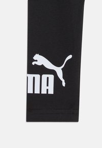 Melni legingi ar baltu Puma logo un tekstu. Stiepjams audums; gluda tekstūra. Standarta dizains, bez papildu rakstiem vai akcentiem.