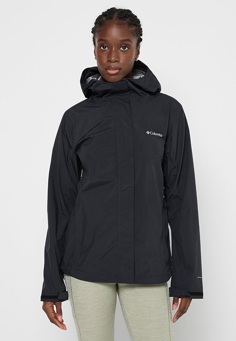 Femme portant une veste de pluie noire à capuche Columbia avec fermeture éclair à l'avant, se tenant contre un fond clair.