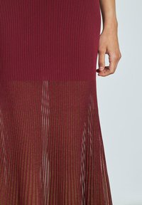 Vestido burdeos de canalé con parte superior ajustada y falda inferior translúcida que presenta plisados verticales y sutiles acentos metálicos.