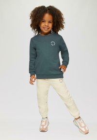 Tumši tirkīza sporta krekls ar apaļu kakla izgriezumu un baltu logotipa dizainu, savienots ar gaiši krēmkrāsas sporta biksēm ar atbilstošu logotipu un pelēki rozā kedām.