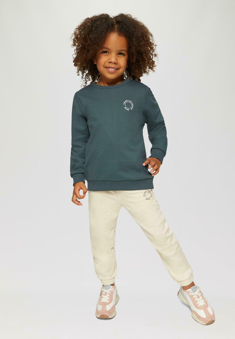 Tumši tirkīza sporta krekls ar apaļu kakla izgriezumu un baltu logotipa dizainu, savienots ar gaiši krēmkrāsas sporta biksēm ar atbilstošu logotipu un pelēki rozā kedām.